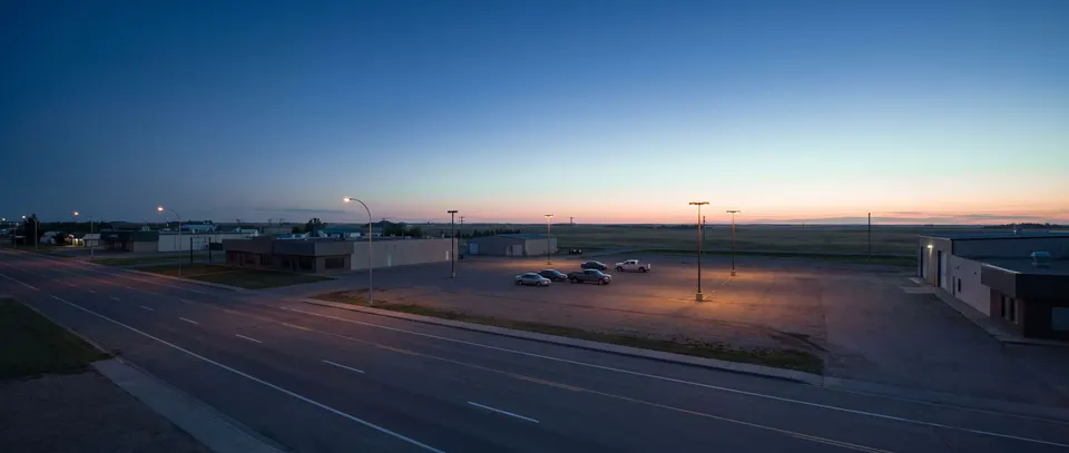 A wide commercial avenue at blue hour on the edge of a prairie city, a parking lot under tall lamps with a few cars, flat land and a pale band of light on the horizon.
